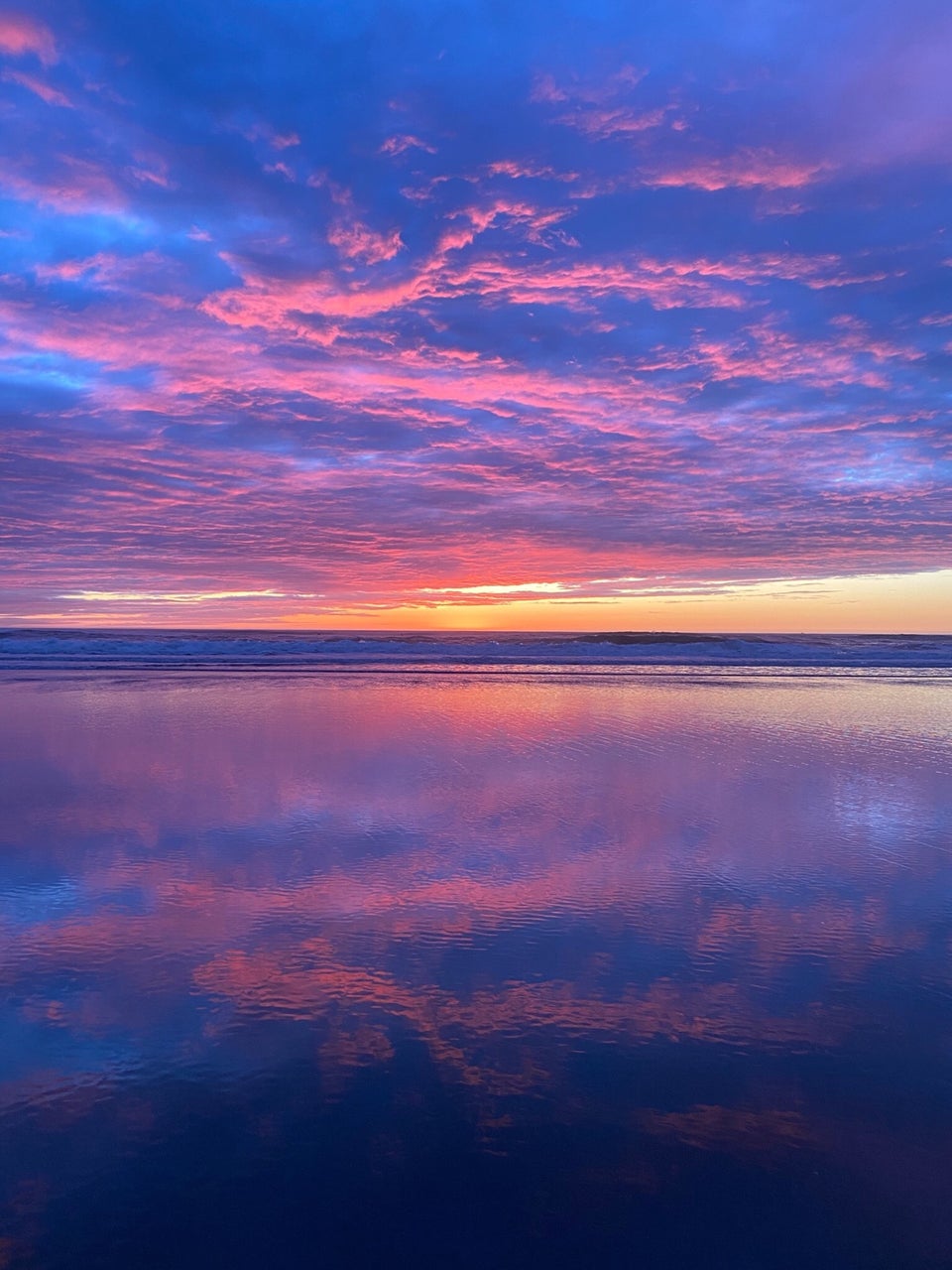 Blue, purple, pink clouds lighting up a post-sunset sky above an orange yellow horizon and distant Pacific Ocean waves crashing on a shore, wet sand in the foreground reflecting the beautiful sky.