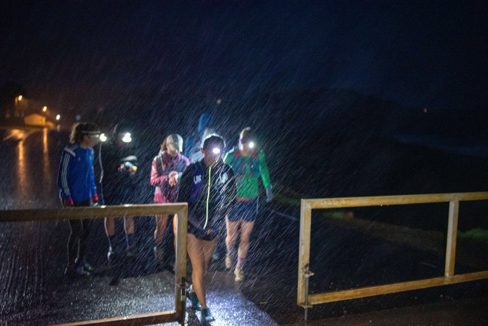 Half a dozen runners wearing headlamps, rain gear, and shorts, starting a run at a gate in the darkness, their headlamps lighting up the pouring rain, Rodeo Beach in the background.