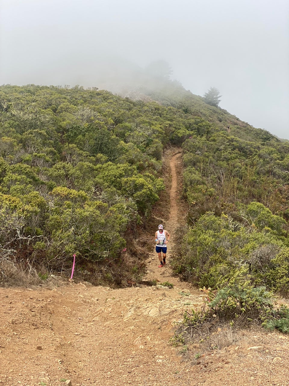 Fogged-in view of a lush green hillside, narrow singletrack trail cutting up from the right and approaching the center, with Tantek power hiking uphill, grinning, water bottle in right hand, holding his side with his left.