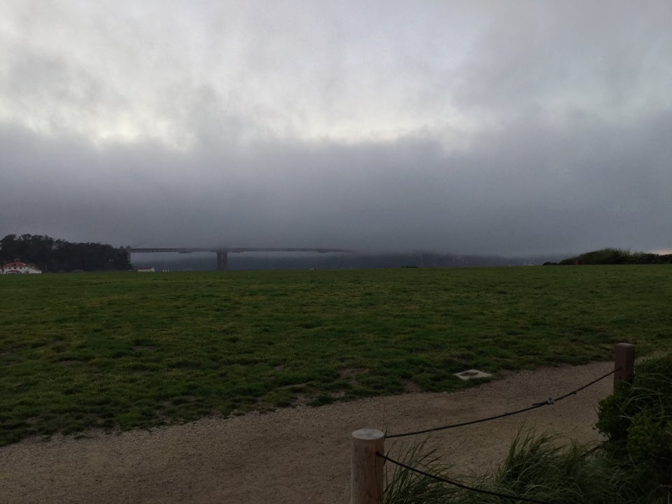 Heavy fog covering all but the bottom edge of the Golden Gate Bridge, above a very green Crissy field in the foreground, with a gravel path immediately in front of that.