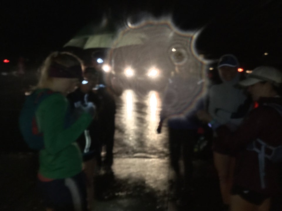Runners clustering in the rainy darkness, Bryan with a green umbrella, backlit by headlamps from cars in the parking lot, also lighting up a drop of water on the camera lens.