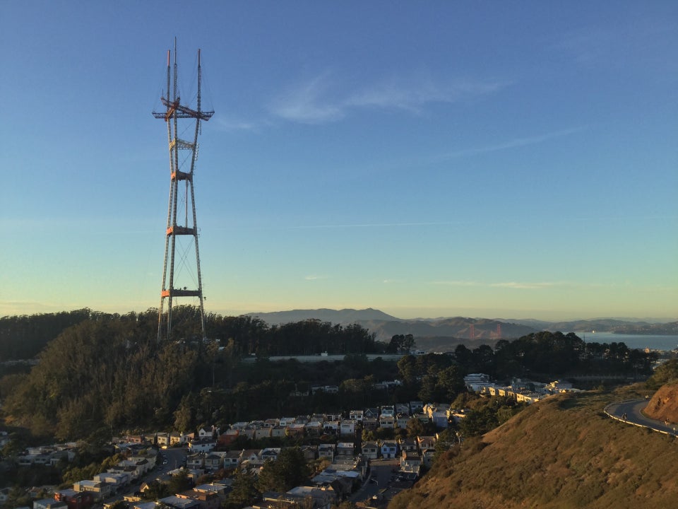 Sutro Tower nearby and a tiny Golden Gate Bridge in the distance, both lit with orange light from the sunset, blue skies with a few small cloud streaks, Mount Tam visible in the distance.