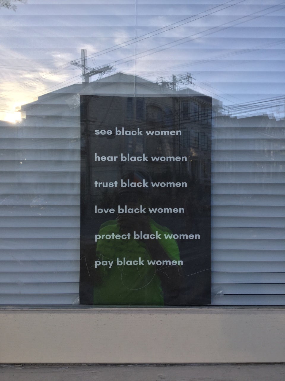 Black sign with white text in a window with white mini-blinds behind it, with six lines: see black women, hear black women, trust black women, love black women, protect black women, pay black women.