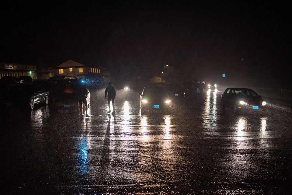 Several cars parked at and a few more approaching the Rodeo Beach parking lot, a couple of buildings in the background lit from inside, silhouettes of a few people backlit by headlamps reflecting on the wet asphalt in the dark.