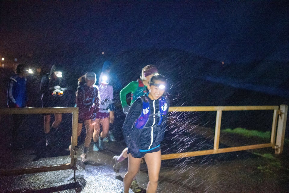 Half a dozen runners wearing headlamps, rain gear, and shorts, starting a run crossing through a gate in the darkness, their headlamps lighting up the pouring rain, Rodeo Beach in the background.