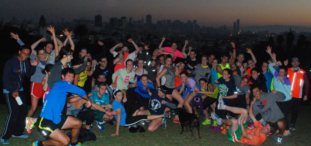 November Project San Francisco group photo at Dolores Park just before sunrise, the barely backlit outline of the downtown skyline visible behind them.